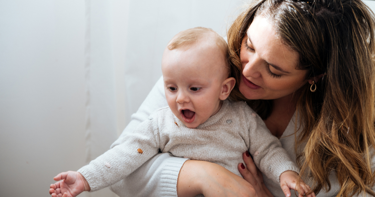 A mother cradles her laughing baby, both with joyful expressions, in a cozy indoor setting with a white background.