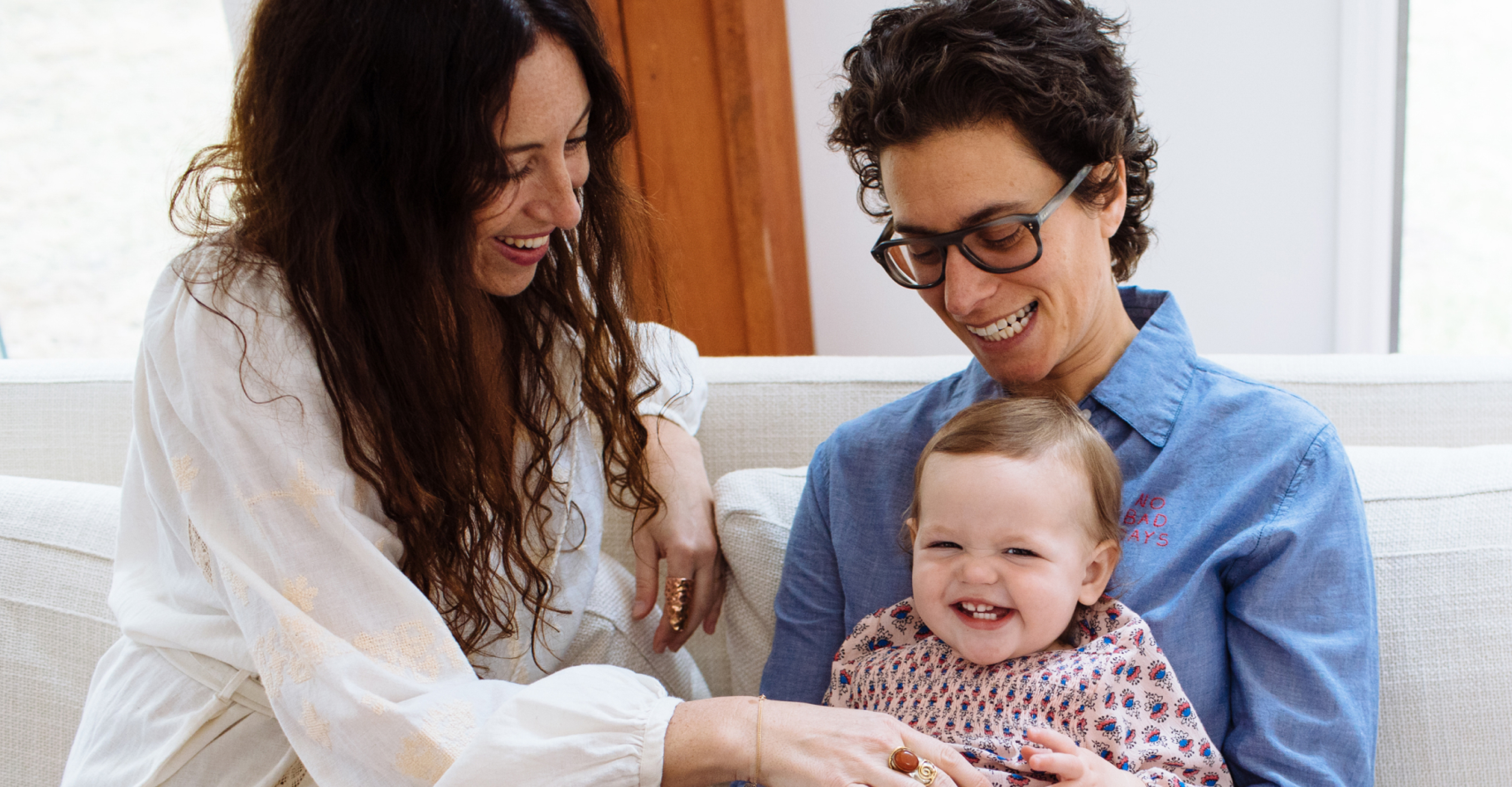 Two women and a laughing baby sitting comfortably on a white sofa, sharing a joyous family moment together.