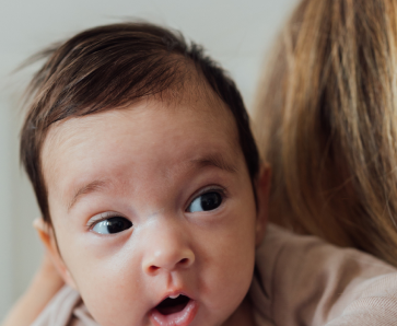 A curious baby with dark hair glancing over a parent's shoulder, with an expression of wonder and big, bright eyes.