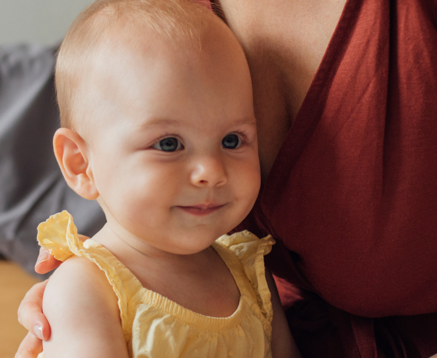 A smiling baby in a yellow outfit held by a woman in a maroon dress, both seated on the floor, sharing a tender moment together.