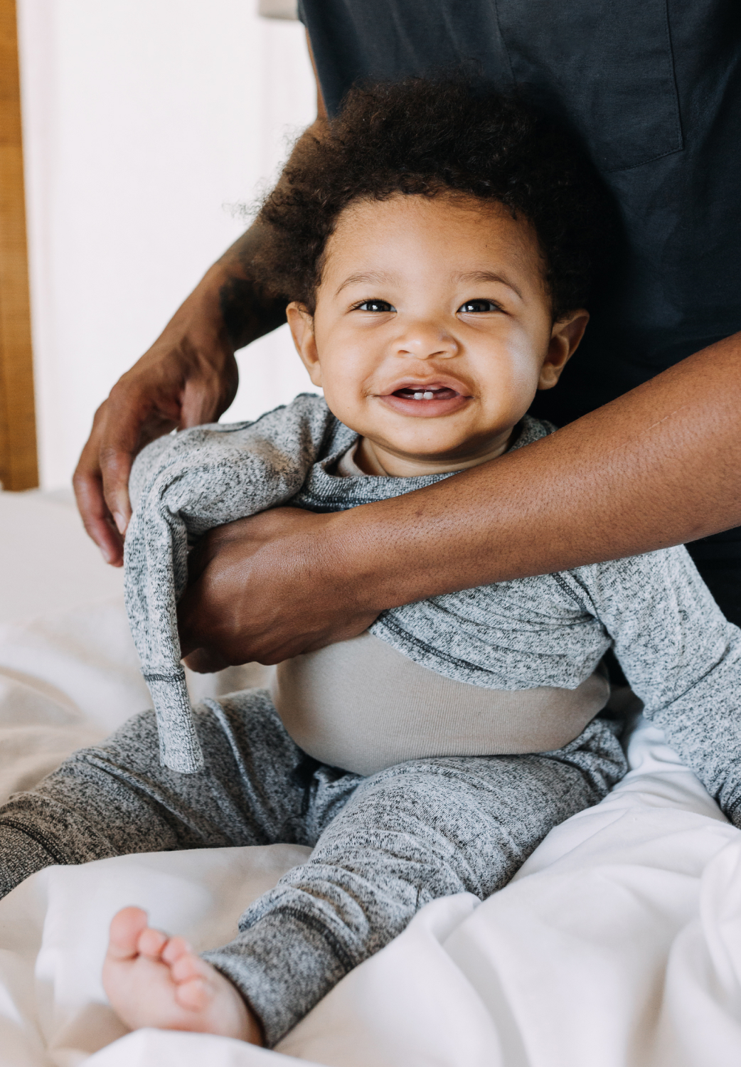 A smiling toddler dressed in gray being lovingly held by his father, who is wearing a dark shirt, in a cozy bedroom setting.